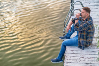 Young man sitting sipping coffee on a wooden jetty or boardwalk at sunset with reflections of the glowing sun on the water