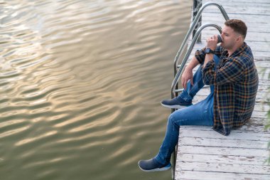 High angle view of a casual guy relaxing in a wooden deck above a tranquil lake at sunset drinking a cup of coffee