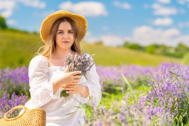 Happy friendly young woman picking fresh lavender in summer standing in the field in fresh white frock and sunhat holding a bunch of flowers