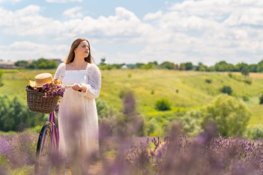 Young woman wheeling her bicycle through farms fields dotted with purple lavender looking off to the side watching
