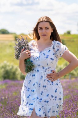 Young woman posing with a bunch of fresh lavender in a field standing with hand in hip looking at the camera