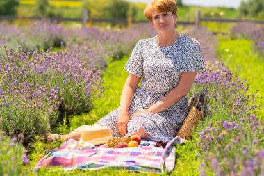 Attractive middle aged woman enjoying a summer picnic sitting on a rug between rows of flowering lavender