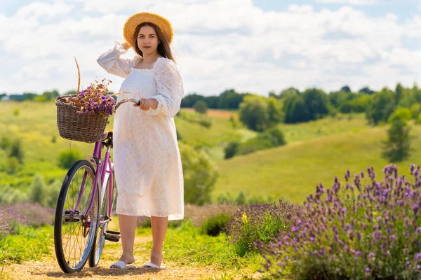 Young woman in a lacey white summer dress pushing her bike filled with fresh lavender through the fields in summer