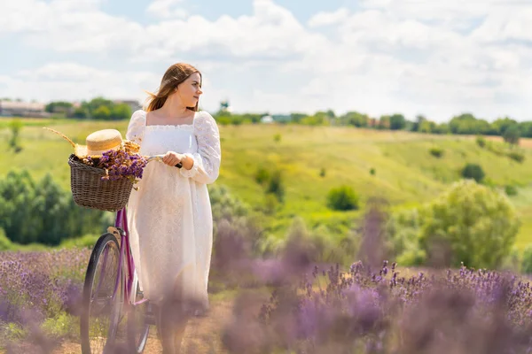 Young country woman wheeling her bicycle through lavender fields with a straw sunhat resting on the handlebars