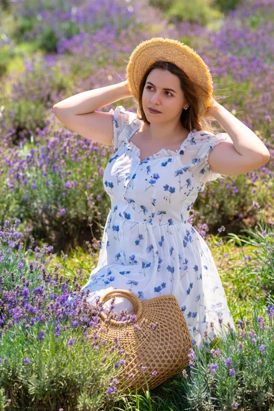 Woman holding her straw sunhat on a breezy day outdoors as she sits in a farm field amongst the lavender