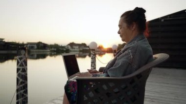 Sunset 'te sakin bir gölün üstündeki güvertede dizüstü bilgisayar kullanan yaşlı bir kadın oturuyor.