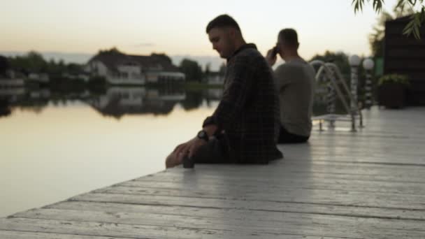静かな湖を見下ろす木製のデッキに座って夕日に2人の男性のソフトフォーカスイメージ コピースペース — ストック動画