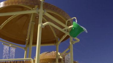 Bir su kaydırağının çatısından akan sular, yazlık bir turizm beldesinde, güneşli mavi gökyüzüne çarpıyor.