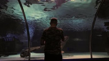 Man viewing an old rusted ships anchor from a shipwreck lying on the sand in a large aquarium tank in a rear view conceptual of vacations and tourism