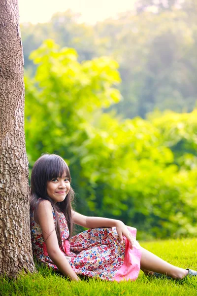 Smiling little asian girl sitting under a tree — Stock Photo, Image