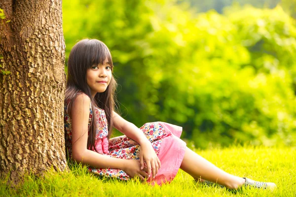 Smiling little asian girl sitting under a tree — Stock Photo, Image