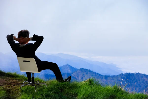 Young businessman who sits on a chair at the top of the mountain — Stock Photo, Image