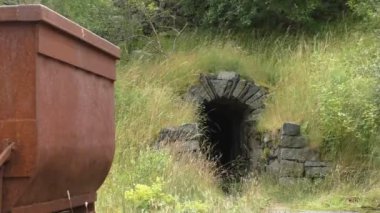 Çocuklu bir grup insan, hepsi kask takıyor, paslı maden arabasının yanından geçiyorlar, turistlerin tur rehberini takip ederek tepedeki eski maden girişine doğru yürüyorlar..