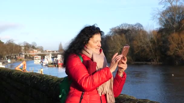 在一个阳光明媚的冬日里 穿着红色夹克 留着长长的黑头发的游客妇女在河边用智能手机拍摄切斯特城的风景 — 图库视频影像