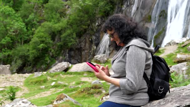 Şelalenin Kenarındaki Kayada Oturan Kadın Cep Telefonundaki Son Fotoğraflara Bakıp — Stok video