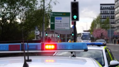 Stockport town street scene. Close up of red emergency light flashing on police car roof, another vehicle reversing to join traffic flowing past, selective focus, blurred background.