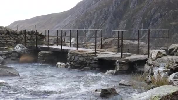 手持相机在大风中拍摄 威尔士山区一座小型人行桥将水滴从溪流表面刮起 — 图库视频影像
