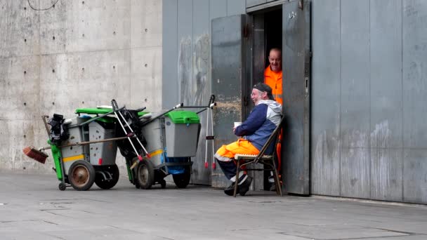 オレンジの制服と安全靴を着て街の清掃員は オープンストレージルームでリラックスしたランチ休憩をしながら話して笑っています — ストック動画