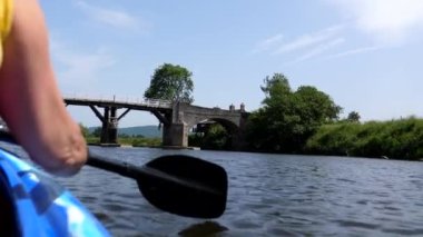 Double kayak rear kayaker view of woman in front paddling on Welsh and English border river Wye at Whitney on Wye village in Herefordshire, England; approaching wooden toll bridge, car crossing.