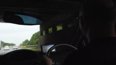 Rear left side view of driver with eyeglasses driving private car on highway in England, using sat nav or GPS, selective focus on man's head.