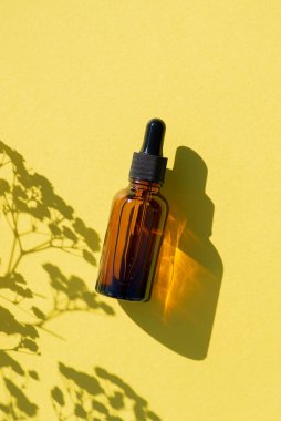Siyah kapaklı kehribar cam damlası şişesi. Bitki gölgesi ile sarı arkaplanda üst görünüm.