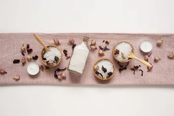 Tigelas de madeira e garrafa cheia de sal marinho banho branco com botões de rosa seca em tecido rosa natural. — Fotografia de Stock