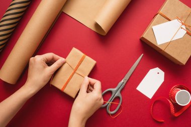 Kraft hediye kutusunda kırmızı arka planda turuncu iplikle fiyonk yapan genç bir kadın. Kutlama için hediye paketleme ve hazırlama. Yılbaşı arkaplanı makas, kağıt ve yazıtlar için etiket ile