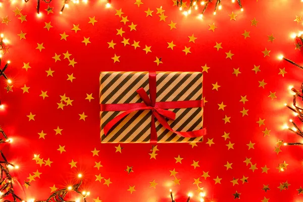 Scatola regalo a strisce Kraft con nastro su fondo rosso con confetti dorati a forma di stelle. — Foto Stock