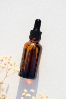 Siyah kapaklı kehribar cam damlası şişesi. Kuru çiçeklerle beyaz arkaplanda üst görünüm.