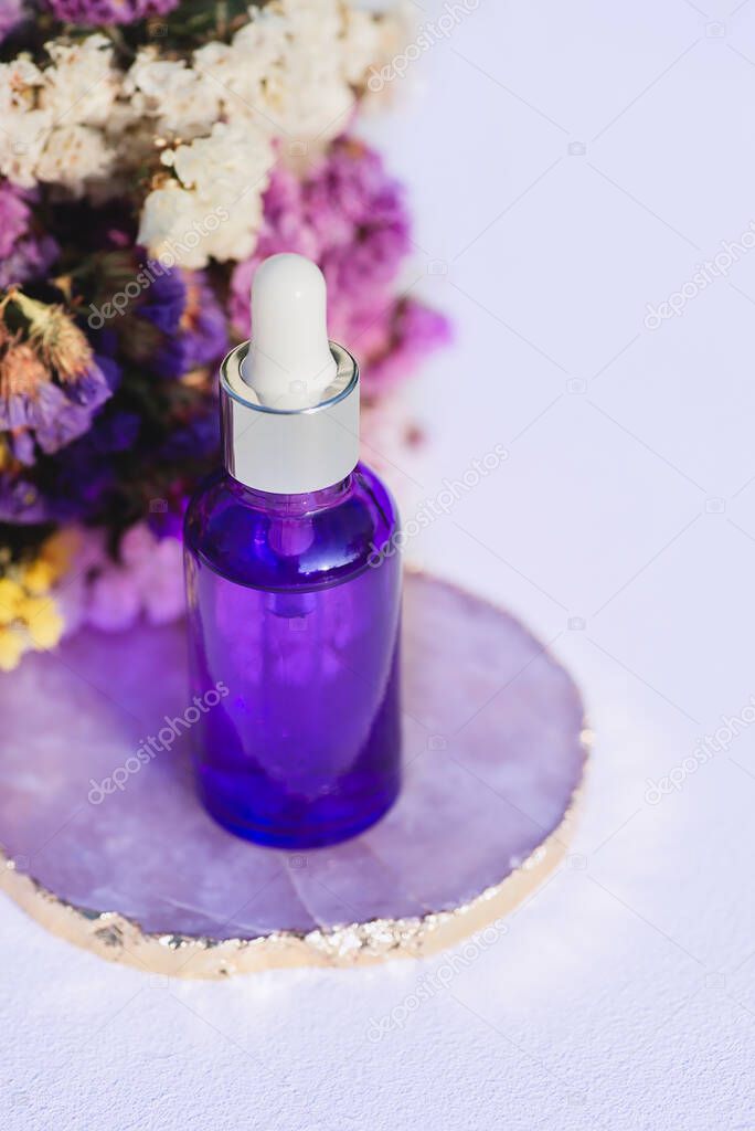 Purple glass dropper bottle on stone podium. White background with dry flowers. Container mockup with cosmetic oil or serum. Showcase for the presentation skincare products.