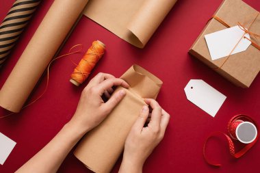 Kırmızı arka planda el işi kağıdına hediye paketleyen genç bir kadın. Kutlama için hediye hazırlığı. Yılbaşı arkaplanı makas, kağıt ve yazıtlar için etiket ile