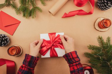 Kırmızı kurdeleli beyaz hediye kutusu Kraft arka planında el ele tutuşuyor. Kutlama için hediye paketleme ve hazırlama. Yılbaşı arkaplanı ağaç süsü ve ışıklarıyla