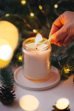 El, yeşil Noel ağacı dalları ve ışıklarının arka planında kibritle bir mum yakar.