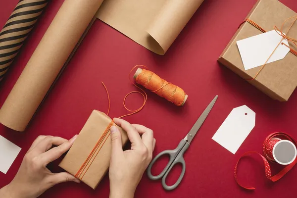 Junge Frau macht Schleife auf Kraft-Geschenkbox mit orangefarbenem Faden auf rotem Hintergrund. — Stockfoto