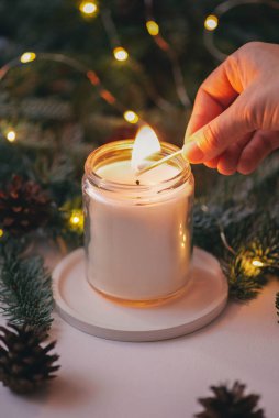 El, yeşil Noel ağacı dalları ve ışıklarının arka planında kibritle bir mum yakar.