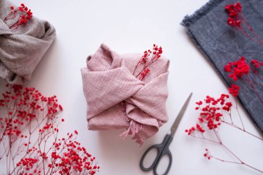 Beyaz arka planda, doğal kumaşlı, makaslı ve kuru çiçekli bir hediye kutusu. Çevre ve sürdürülebilir hediye paketleme fikirleri. Furoshiki ambalajlı şenlik arkaplanı. Geri dönüşüm ve çevre dostu alternatifler.
