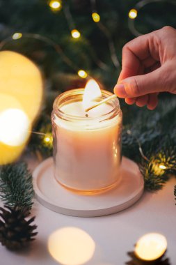 El, yeşil Noel ağacı dalları ve ışıklarının arka planında kibritle bir mum yakar.