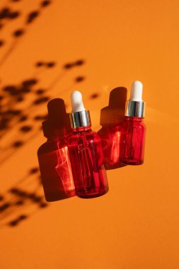 Turuncu arka planda gümüş kapaklı kırmızı cam damlalar. Güzel gün ışığında cilt bakım ürünleri. Doğal bitki gölgesi ile doğal kozmetik konsepti. Yüz ve vücut bakımı için güzellik kavramı.