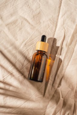 Kehribar cam şişelerde damla pipeti serumla ya da açık gri kumaş arka planında gerekli yağla. Cilt bakımı kozmetik sunumu, gün ışığı ve palmiye yaprakları gölgesi. Yüz bakımı için güzellik konsepti