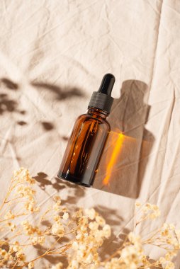 Kehribar cam şişe, serumlu damlatan pipet ya da açık gri kumaş arka planında gerekli yağ. Güneş ışığı ve kuru çiçeklerle cilt bakımı kozmetik sunumu. Yüz bakımı için güzellik konsepti