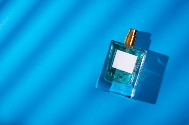 Mavi arka planda etiketi olan saydam bir parfüm şişesi. Gün ışığıyla güzel kokulu bir sunum. Doğal malzemelerdeki güzel gölgeli moda konsept. Kadın ve erkeğin özü..