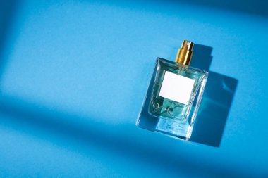 Mavi arka planda etiketi olan saydam bir parfüm şişesi. Gün ışığıyla güzel kokulu bir sunum. Doğal malzemelerdeki güzel gölgeli moda konsept. Kadın ve erkeğin özü..