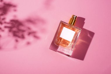 Pembe arka planda etiketi olan saydam bir parfüm şişesi. Gün ışığıyla güzel kokulu bir sunum. Çiçekli gölgeli doğal malzemelerde moda olan bir kavram. Kadın özü..