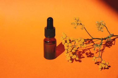 Damla pipetli bir şişe serum ya da gerekli yağ. Turuncu arka plan, gün ışığı ve sarı çiçekler. Cilt bakım ürünleri, doğal kozmetik ürünleri. Yüz ve vücut bakımı için güzellik konsepti