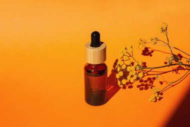 Damla pipetli bir şişe serum ya da gerekli yağ. Turuncu arka plan, gün ışığı ve sarı çiçekler. Cilt bakım ürünleri, doğal kozmetik ürünleri. Yüz ve vücut bakımı için güzellik konsepti