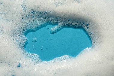 Mavi arka planda köpük. Köpüklü sabunlu sıvı doku. Doğal güneş ve gölgeler. Cilt temizleme kozmetiği üst görünümde.