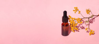 Damla pipetli bir şişe serum ya da gerekli yağ. Pembe arka plan, gün ışığı ve sarı çiçekler. Cilt bakım ürünleri, doğal kozmetik ürünleri. Yüz ve vücut bakımı için güzellik konsepti