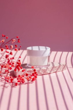 White jar of cosmetic on pink concrete podium for product presentation. Skin care cosmetic with beautiful shadows with red flowers. Beauty concept for face body care.