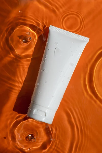 Plastic white tube for cream or lotion on beautiful bursts and glare. Skincare or sunscreen cosmetic on orange background. Beauty concept for face care. Natural sunlight and water spills. Summer mood.