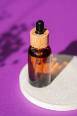 Podyumda siyah kapaklı kehribar cam damlası şişesi ve mor arka planda bitkilerin gölgesi. Cilt bakım ürünleri, doğal kozmetik ürünleri. Yüz ve vücut bakımı için güzellik konsepti